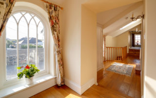 A stunning large ornate window letting light into a spacious hallway with wooden floors and sloping ceiling